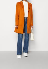 Abrigo de mezcla de lana en color naranja, de un solo botón con solapas de muesca, llevado sobre un suéter de cuello alto blanco. Combinado con jeans de tiro alto azules y zapatillas blancas.