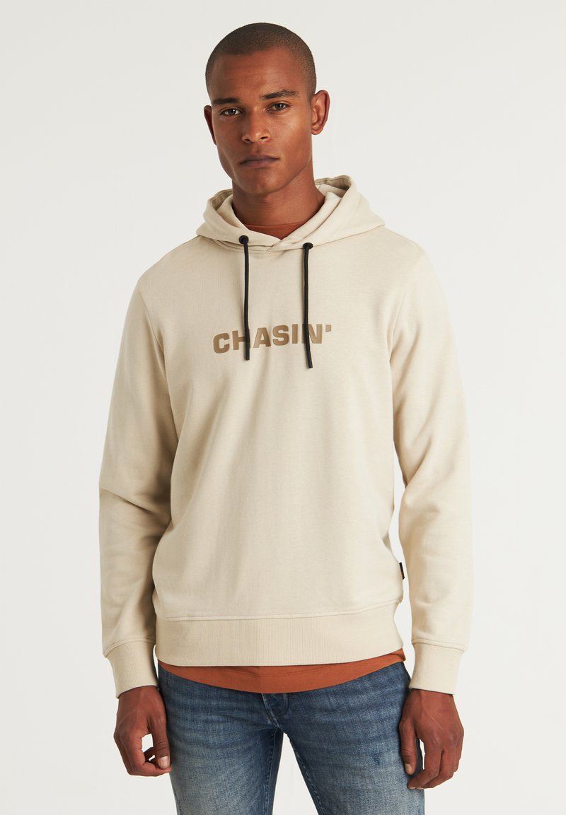 Beige kapucnis pulóver fekete zsinórokkal. "CHASIN'" felirat barna színben a mellkason. Hosszú ujjú, bordázott mandzsettával, alatta rozsdaszínű pólóval.