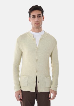 Uomo che indossa un cardigan lavorato a maglia color crema con bottoni sopra una camicia bianca e pantaloni scuri, in piedi contro uno sfondo chiaro e semplice.