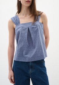 Modra tkanina bluzica s kvadratnim izrezom, širokimi naramnicami in naborki spredaj. Tkanina ima subtilno teksturo in vzorec, v kombinaciji z visokim pasom denim hlačami.