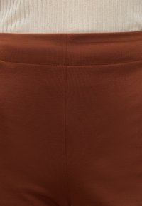 Gros plan d'une personne portant un pantalon marron taille haute et un pull côtelé crème rentré.