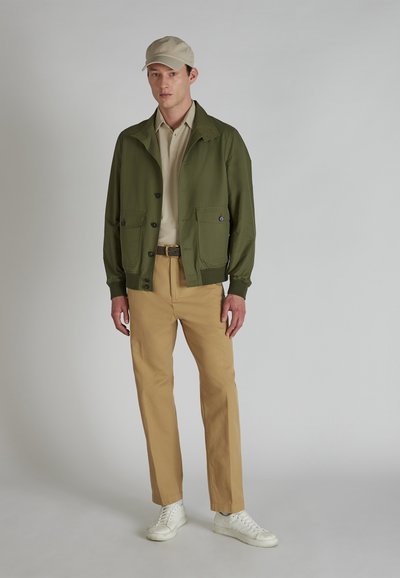 Olijfgroene jas met twee voorzakken, lichtbeige polo, tan broek en witte sneakers. Model draagt een lichtbeige pet.