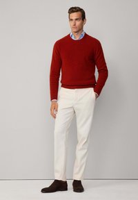 Roter Wollpullover mit Rundhalsausschnitt, kombiniert mit weißen Hosen. Das Model trägt dunkelbraune Wildlederschuhe. Einfaches, klares Design und Passform.