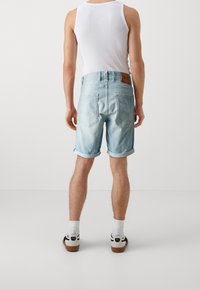 Pantalones cortos de mezclilla azul claro con puños enrollados, dos bolsillos traseros, cintura de altura media y una etiqueta de cuero marrón. Combinados con una camiseta sin mangas blanca y zapatillas.