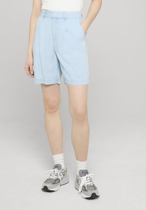 Person som har på seg lyseblå shorts til kneet, hvite sokker, grå og hvite joggesko, og en hvit topp, står med en hånd i lommen.