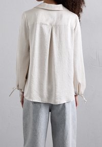 Blusa bianca accartocciata con colletto, schiena plissettata e maniche elastiche legate con stringhe, abbinata a jeans a vita alta di un blu chiaro.