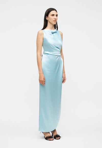Γυναίκα με μακριά σκούρα μαλλιά, που φοράει ένα αμάνικο, ανοιχτό μπλε σατέν βραδινό φόρεμα και μαύρα ψηλοτάκουνες σανδάλια με φτερά, στέκεται σε λευκό φόντο.