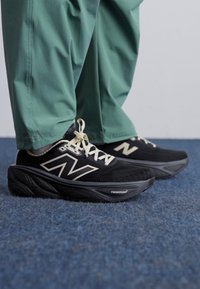 Baskets noires avec une tige texturée, logo et lacets blancs, et une semelle noire rembourrée. Portées avec un pantalon vert ample.