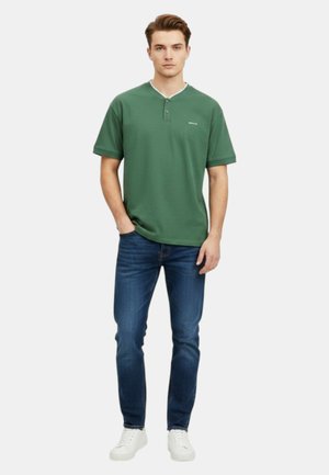 Giovane uomo in piedi che indossa una maglietta henley verde a maniche corte, jeans slim fit blu e sneakers bianche su uno sfondo semplice.