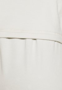 Tela de un beige claro que presenta un detalle de cintura fruncida y costuras en contraste a lo largo del borde superior. Textura suave con un acabado liso.
