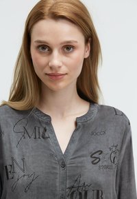 Junge Frau mit langen, hellbraunen Haaren, die ein graues, geknöpftes Hemd mit schwarzem Drucktext und dekorativen Strasssteinen trägt.