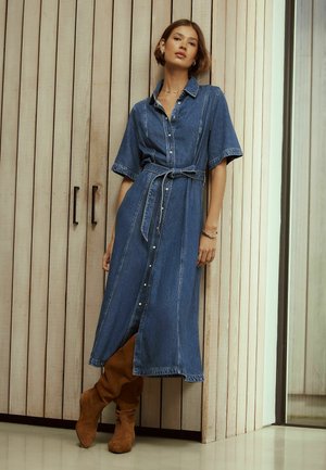 Nő egy fa falnak dőlve, övvel ellátott farmer midi ruhát visel, valamint világosbarna színű, térd fölé érő, velúr csizmát, rövid hajjal és minimális ékszerekkel.