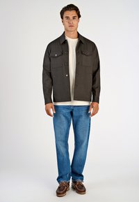 Donkergrijze jack met twee borstzakken, knoopsluiting, gedragen boven een lichte shirt en blauwe jeans. Bruine schoenen met veters maken de outfit compleet.