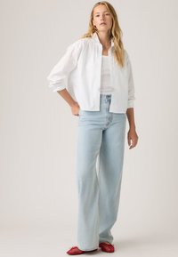 Γυναίκα που στέκεται φορώντας άσπρο πουκάμισο με κουμπιά πάνω από λευκό τοπ, ανοιχτό μπλε τζιν παντελόνι με φαρδιά μπατζάκια και κόκκινα επίπεδα παπούτσια.