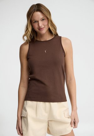 Femme aux cheveux blonds ondulés portant un haut marron sans manches et un short cargo beige, debout devant un fond blanc uni.