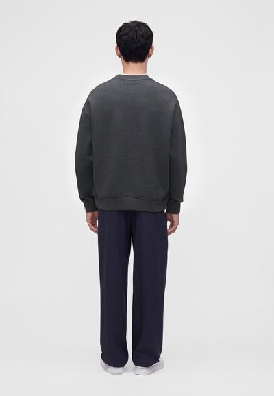 Homme debout, dos à la caméra, portant un sweat-shirt gris foncé, un pantalon large bleu marine et des baskets blanches sur un fond clair uni.
