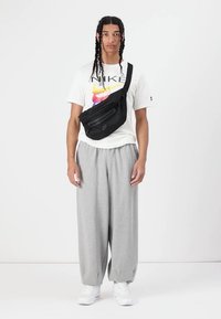 Črn najlonov crossbody nahrbtnik z sprednjim žepom na zadrgo in detajlom logotipa. Model nosi belo grafično majico in sivo trenirka.