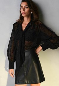 Blusa nera trasparente con pois, collo annodato e maniche lunghe a sbuffo abbinata a una mini gonna in pelle nera con una cucitura anteriore.