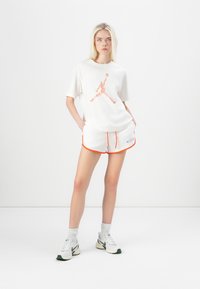 White cotton t-shirt with floral Jordan logo. White shorts with orange trim and logo. Model wears white and green sneakers and white socks.