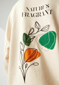 Kremowa tkanina zdobiona haftowanym wzorem w zielone, pomarańczowe i fakturowane akcenty oraz napis „NATURE'S FRAGRANT”.