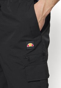 Schwarze Cargo-Hosen mit Seitentaschen, Reißverschluss und besticktem Logo-Patch in Rot, Gelb und Weiß. Aus strapazierfähigem Material gefertigt.