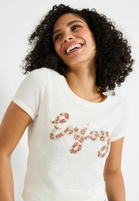 Camiseta blanca de mangas cortas hecha de tela ligera, con un gráfico colorido que dice "Enjoy" en un patrón de tonos rojos, marrones y beiges.