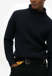 Gestreifter navyblauer Rollkragenpullover mit einem strukturierten Muster. Mit gerippten Bündchen und einer figurschmeichelnden Silhouette, kombiniert mit dunklen Jeans und einem gelben Hemd darunter.