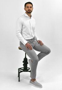Witte button-up shirt met kraag, grijze slim-fit broek en lichtgrijze instapschoenen, zittend op een houten stoel tegen een effen achtergrond.