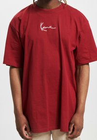 Rotes T-Shirt mit kurzen Ärmeln aus Baumwolle, mit einem weißen Signaturlogo auf der Vorderseite. Entspannter Schnitt mit rundem Ausschnitt und glatter Textur.