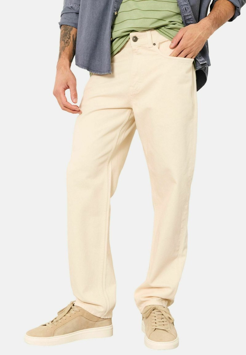 Pantalon en coton couleur crème avec une coupe droite, doté de poches avant, passants pour ceinture et fermeture par bouton. Associé à des baskets beige.
