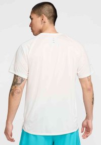 Weißes kurzärmliges Sportshirt mit glatter Textur, rundem Halsausschnitt und kleinen Schlitzen hinten, kombiniert mit blauen Shorts.