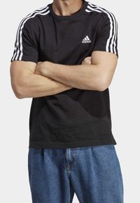 T-shirt nera a maniche corte con design di tre strisce bianche sulle spalle e un piccolo logo bianco sul petto. Materiale in cotone, vestibilità standard.