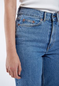 Högmidjade blå denimjeans med en lätt blekning, med kopparnitar, två sidofickor och en texturerad yta.
