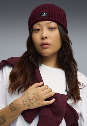 Bordeaux gebreide beanie met een zwart Puma-label, gedragen met een wit shirt en een bordeaux sweatshirt over de schouder gedrapeerd. Hand met zichtbare tatoeages.