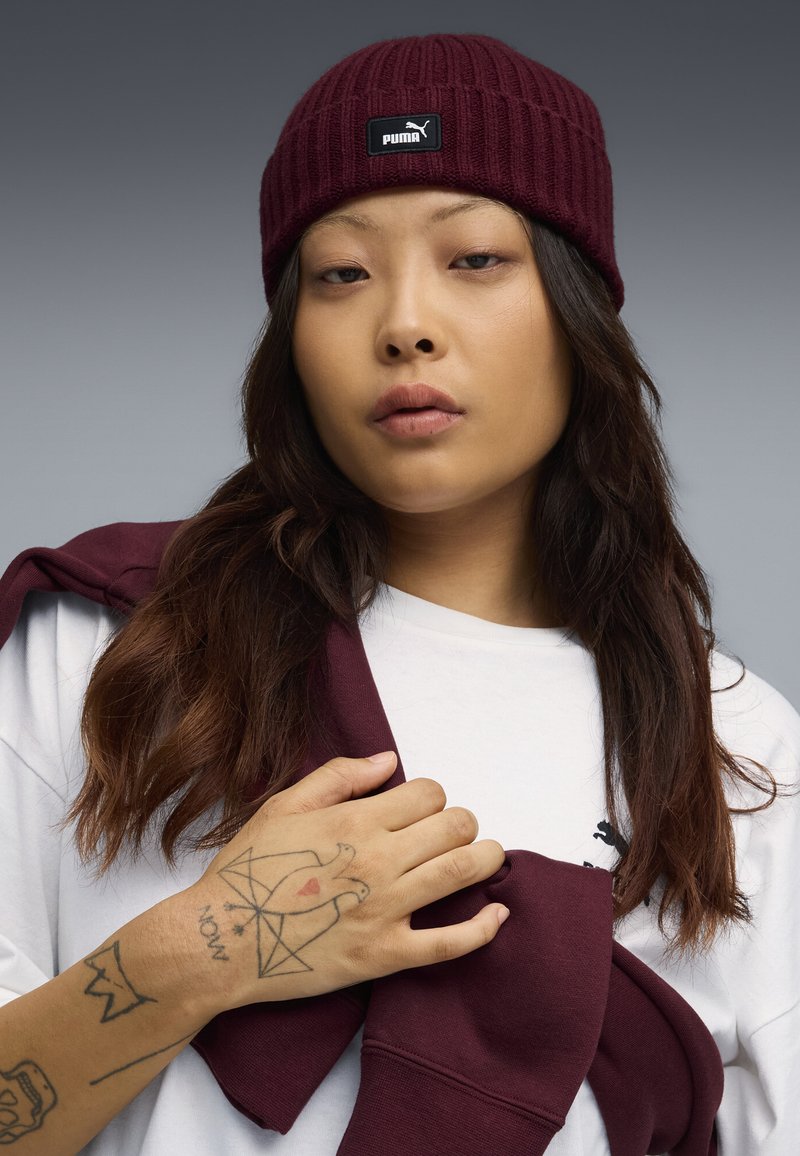 Bordeaux gebreide beanie met een zwart Puma-label, gedragen met een wit shirt en een bordeaux sweatshirt over de schouder gedrapeerd. Hand met zichtbare tatoeages.