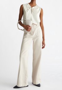 Cremefarvet ærmeløs top med knude foran, parret med højtaljede beige vidde bukser og sorte spidse sko. Bærer en lys håndtaske.