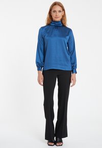 Blaue Satin-Bluse mit hohem Kragen und langen Ärmeln sowie gerafftem Detail am Kragen, kombiniert mit schwarzen Schlaghosen und schwarzen Sandalen mit offenem Zehenbereich.