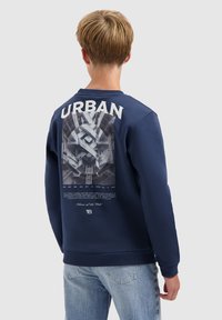 Tiener met kort blond haar, gekleed in een marineblauw sweatshirt met "URBAN" en een architectonische grafiek op de achterkant, in combinatie met lichtblauwe jeans.