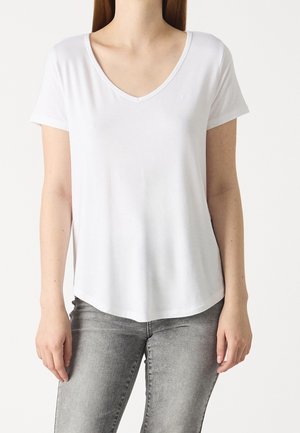 Kvinde iført en enkel hvid V-hals kortærmet T-shirt og falmede grå jeans, stående mod en hvid baggrund.