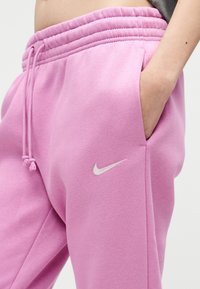 Άτομο που φοράει ροζ φόρμες Nike με το χέρι στην τσέπη, δείχνοντας τη ελαστική μέση και το κεντημένο λευκό λογότυπο Nike swoosh.