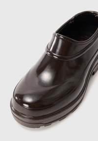 Scarpa nera lucida senza lacci con punta arrotondata, tacco basso, texture liscia e una sottile fascia rialzata attorno al colletto.