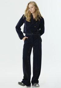 Marineblauwe cropped dubbele-breasted jas met voorzakken, gecombineerd met high-waisted wijde broek. Beide stukken hebben een gladde textuur.