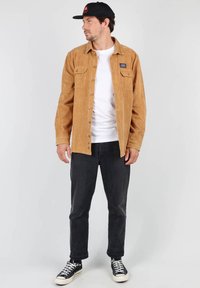 Chemise en velours côtelé marron à boutons sur le devant, avec deux poches poitrine, portée sur un t-shirt blanc, associée à un jean noir et des baskets noires.