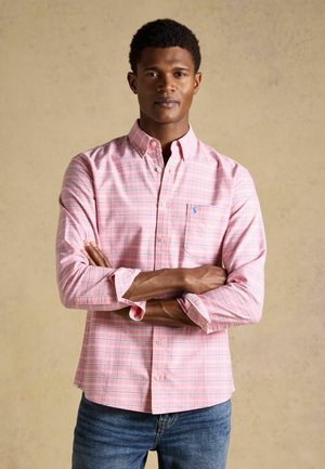 Junger Mann mit verschränkten Armen, der ein rosa kariertes Hemd mit Knöpfen und blaue Jeans vor einem einfarbigen beigen Hintergrund trägt.