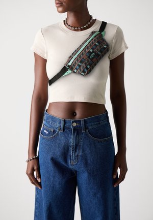 Fanny packki, jossa on monivärisiä geometrisia kuvioita, vetoketjusuljenta ja säädettävä musta hihna, käytetään vaaleanbeigen istuvan paidan ja korkeavyötäröisten farkkujen päällä.