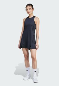 Sleeveless dress in navy fabric with a smooth texture; features side slits and a simple design. Worn with white athletic socks and shoes.