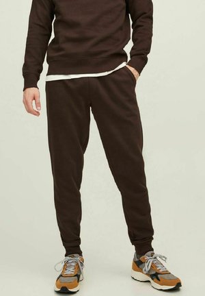 Mann in dunkelbraunen Jogginghosen, passendem Sweatshirt, weißem Unterhemd und orange-grauen, schwarzen Sneakers, der mit einer Hand in der Tasche steht.