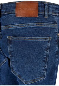 Temno modri traper hlače z rjavim usnjenim oznako "2Y PREMIUM," imajo zadnji žep in kontrastno rumeno šivanje.