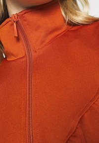 Chaqueta de cremallera de tejido naranja brillante, con un alto cuello, superficie texturizada y detalles de costura visibles a lo largo de la cremallera.