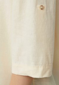 Chemise en lin beige clair avec des manches retroussées et un bouton clair. Le tissu présente une finition douce et texturée.
